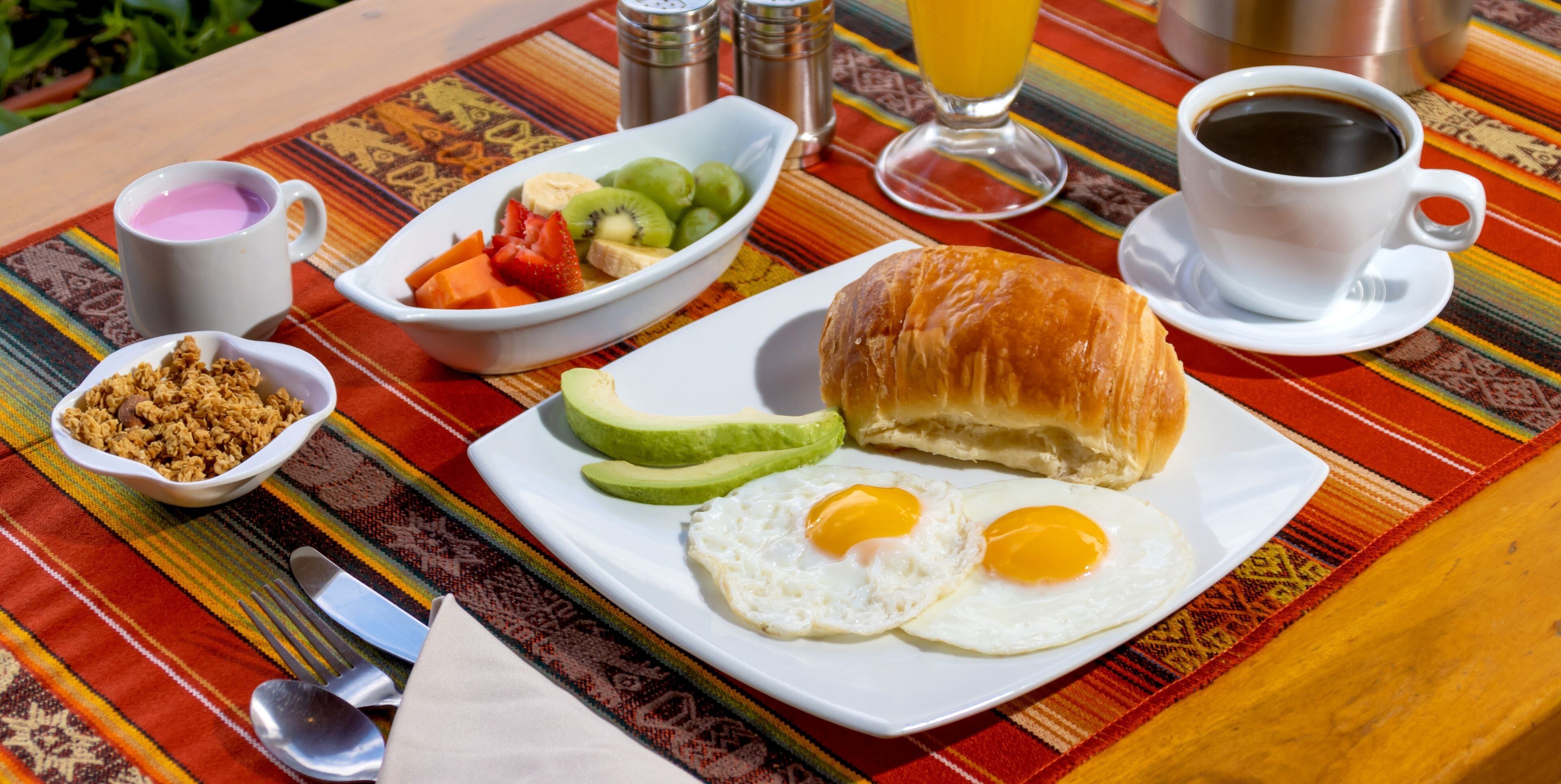 Una mesa de desayuno con un plato de huevos, aguacate y un croissant, un tazón de cereal, una taza de café y un vaso de jugo de naranja.