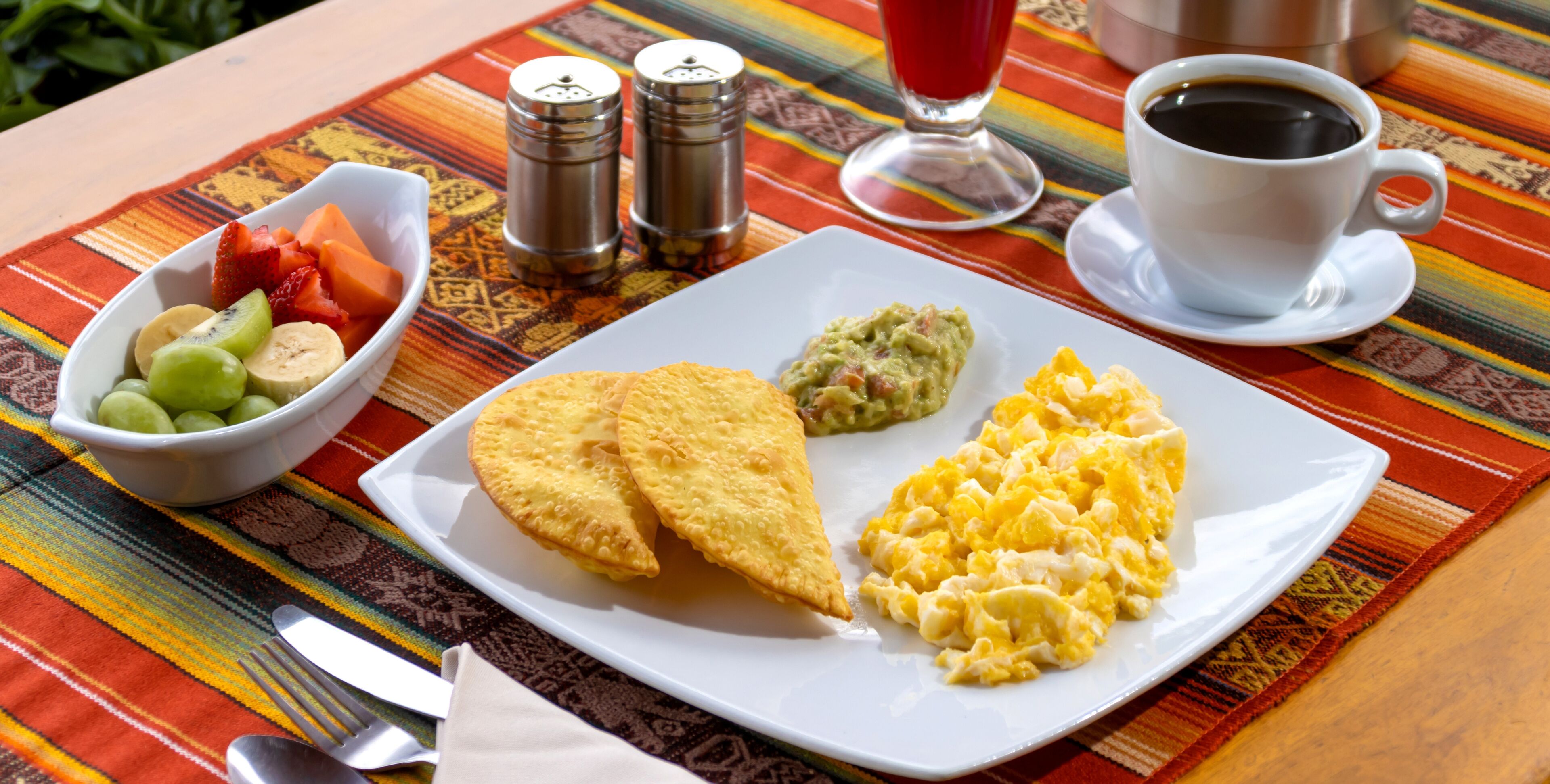 A plate with scrambled eggs, guacamole, and a fried item, possibly a quesadilla, on a colorful tablecloth.