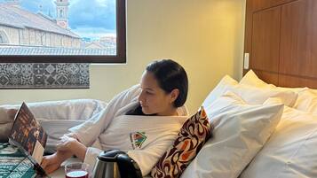 Una persona acostada en la cama con un ordenador portátil, un vaso de bebida roja y una cafetera sobre una bandeja.