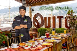 Un chef frente a una mesa de desayuno con vista a la ciudad, un gran letrero con la palabra 'QUITO' y una variedad de alimentos para el desayuno.