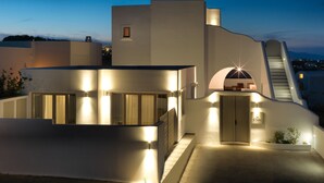 A modern building with a unique architectural design, featuring a curved facade, a staircase leading to an upper level, and a well-lit entrance. - Drops villas (Santorini)