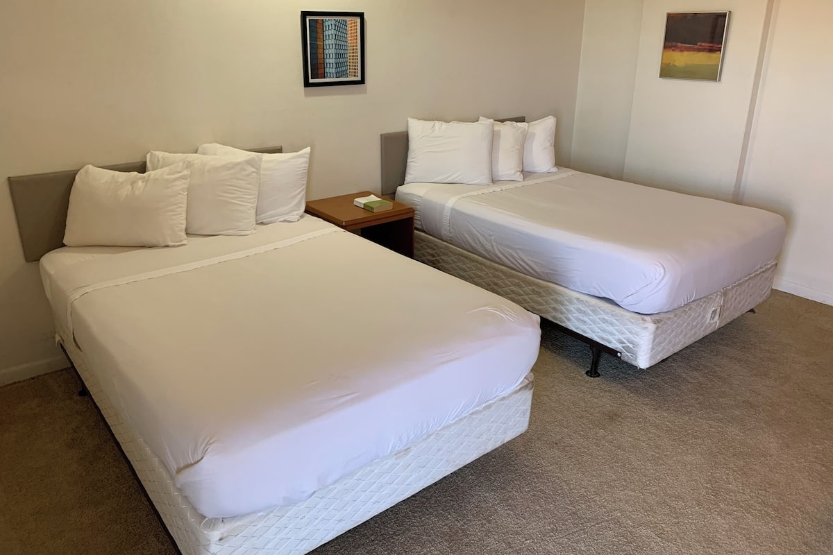 Quarto de hotel com duas camas de solteiro, uma mesinha de cabeceira, um quadro na parede e piso coberto por carpete.
