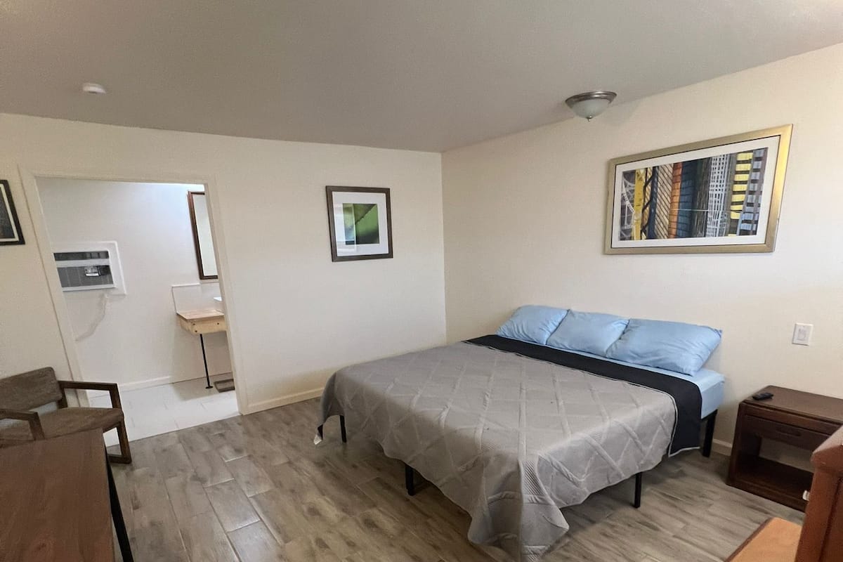 Quarto com cama, escrivaninha e cadeira. Há uma janela com persianas, um quadro na parede e uma porta para outro cômodo.