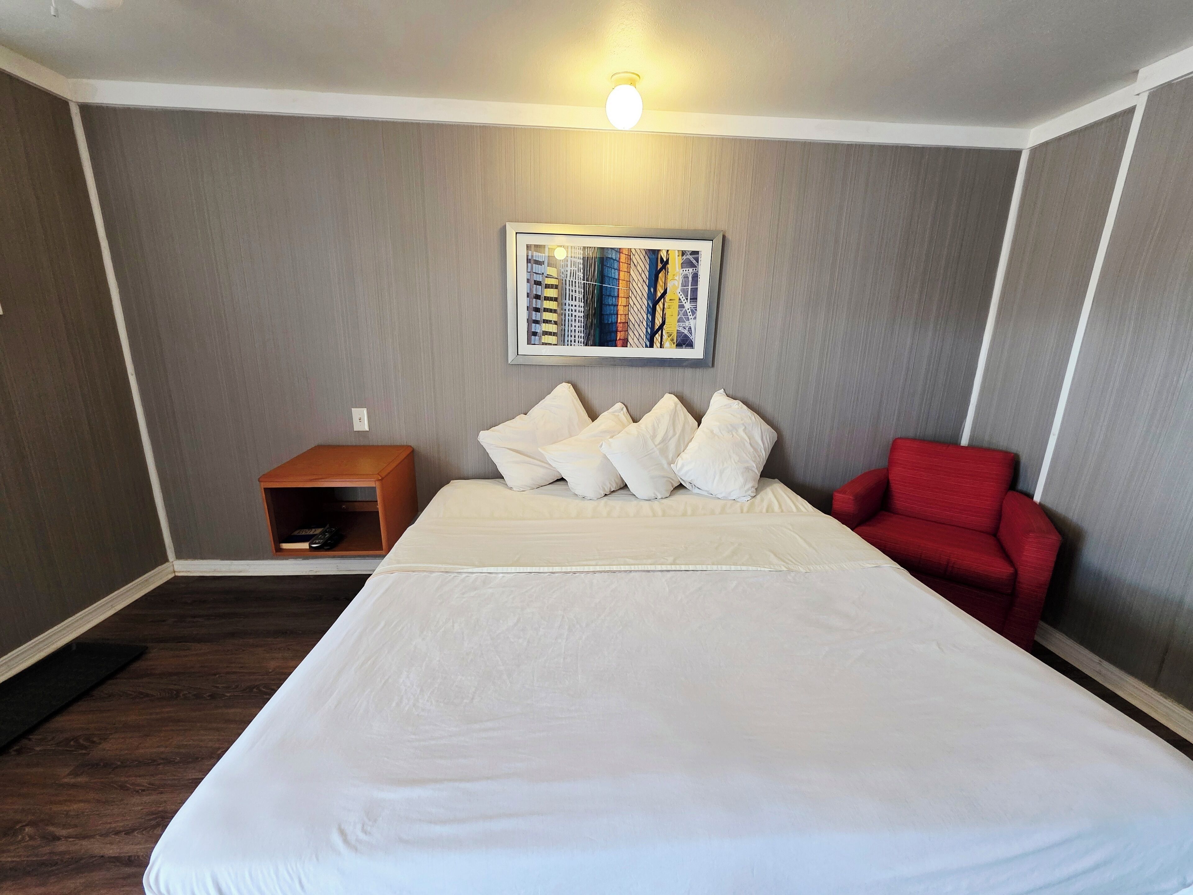 A hotel room with a large bed, a red armchair, a small wooden side table, and a framed picture on the wall.