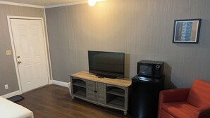 A room with a wooden entertainment center, a television, a microwave, a red chair, and a bed with white bedding. - Sawmill Lodges Motel (Hennessey)