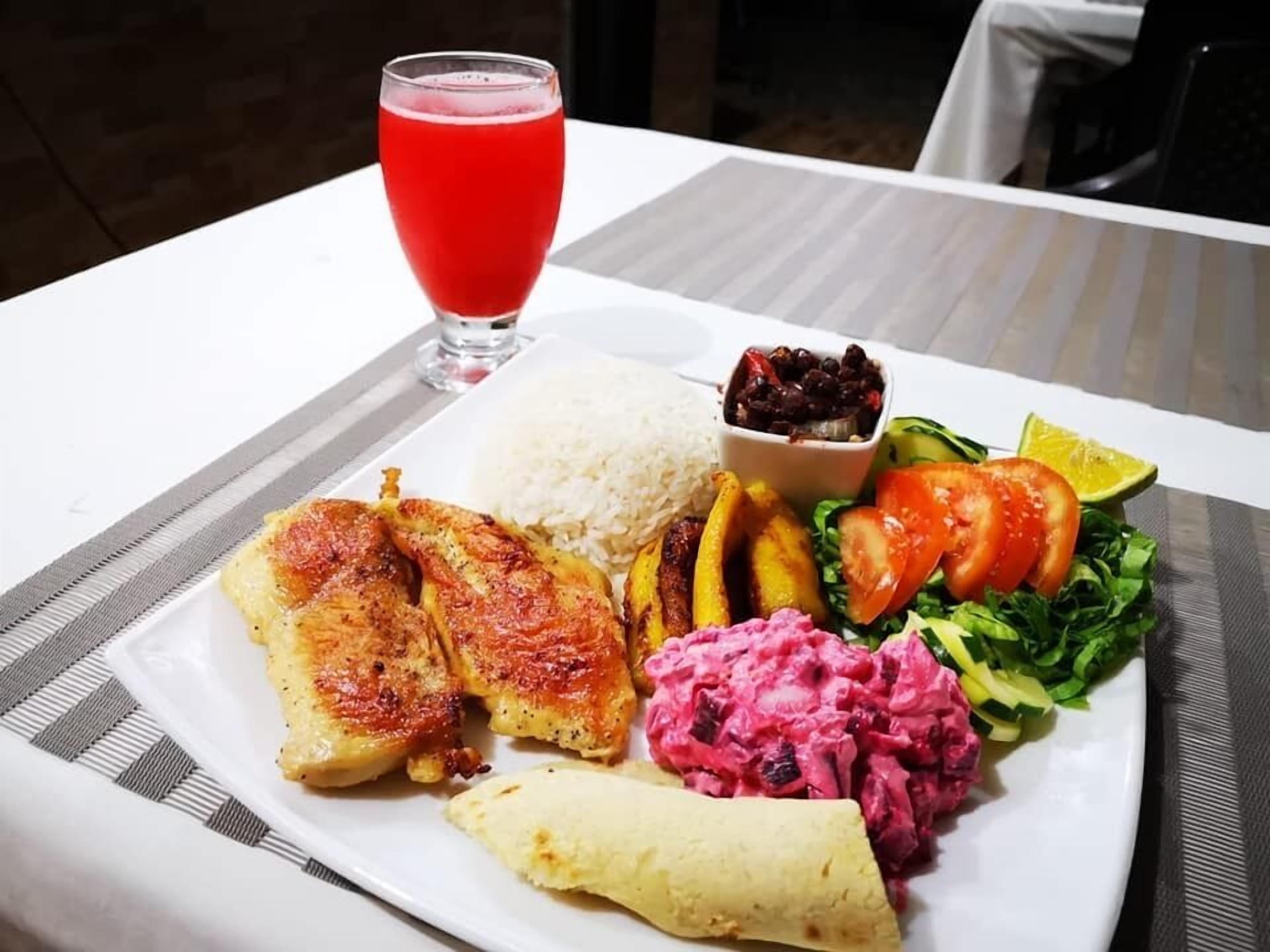 Ein Teller mit gegrilltem Hähnchen, Reis, schwarzen Bohnen, Kochbananen, einem Salat und einem Glas rotem Getränk.