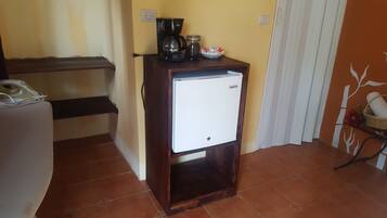 A compact kitchen area with a coffee maker, a small refrigerator, and a shelf with kitchen items.