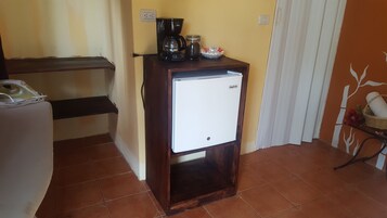 A compact kitchen area with a coffee maker, a small refrigerator, and a shelf with kitchen items.