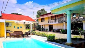 A pool area with a clear blue pool, a covered seating area with chairs, and a building with a red roof and yellow walls.