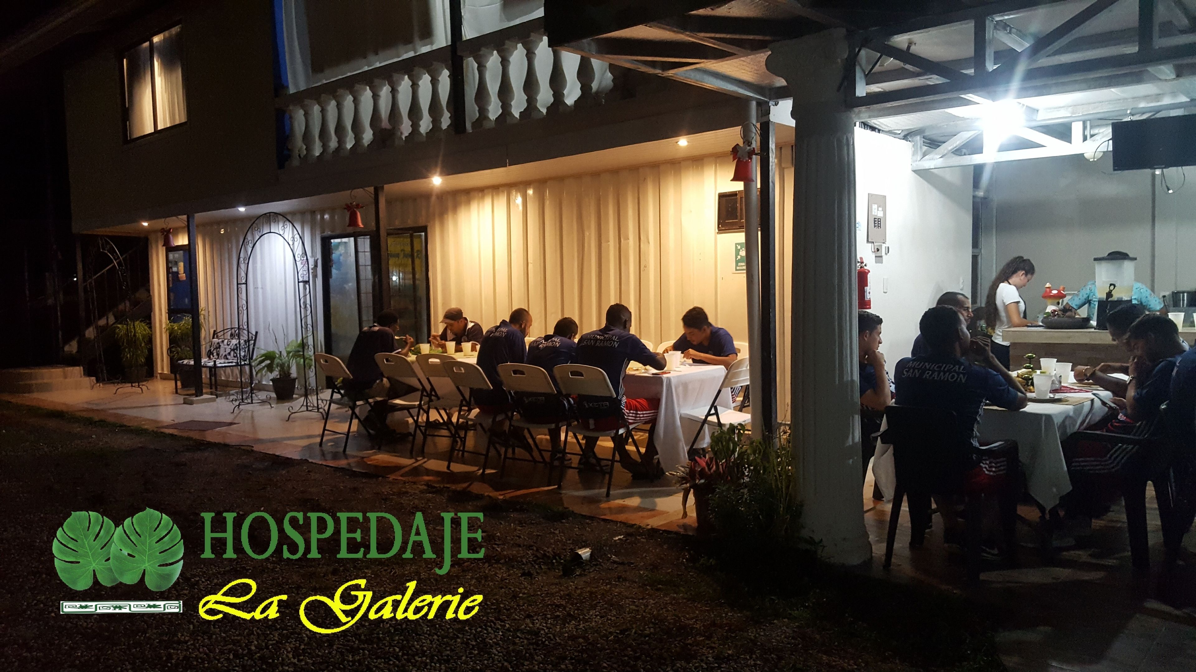 An outdoor dining area with tables and chairs, a covered section with a counter, and a building with a balcony.
