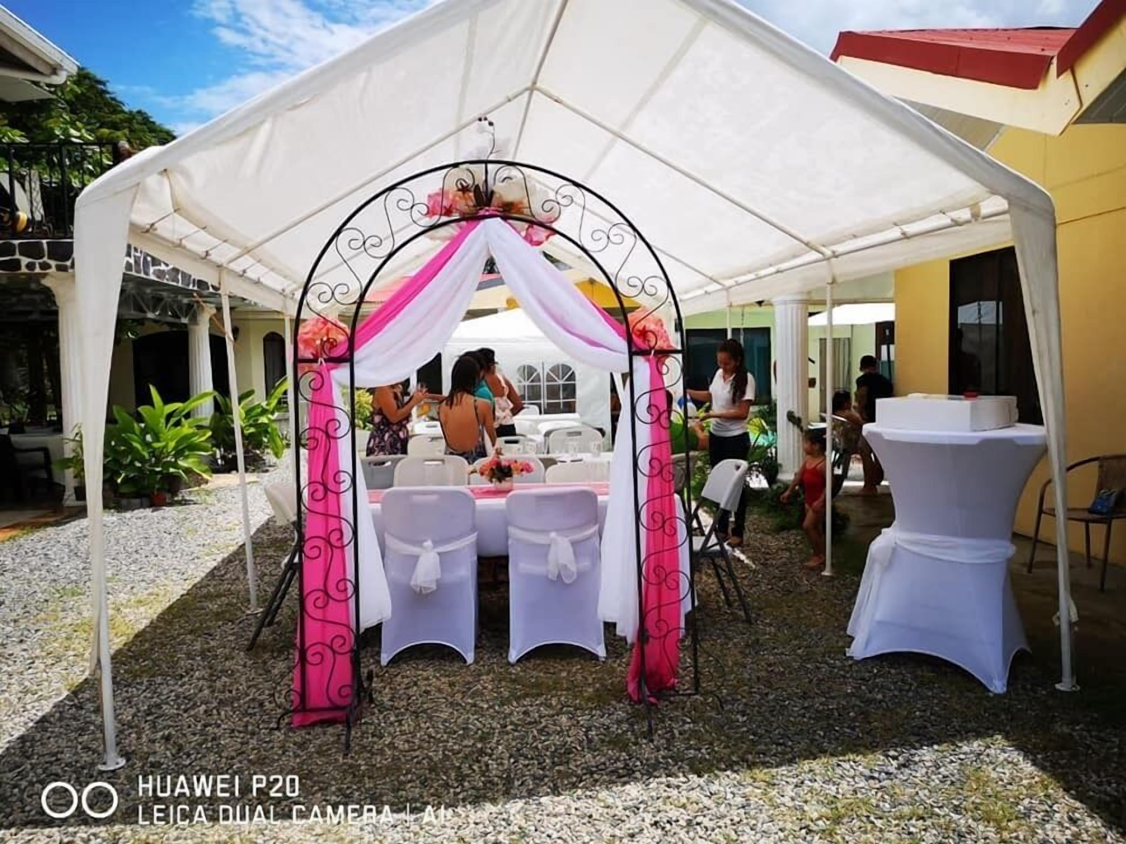 A tented outdoor area with tables and chairs, surrounded by a gravel path and greenery.