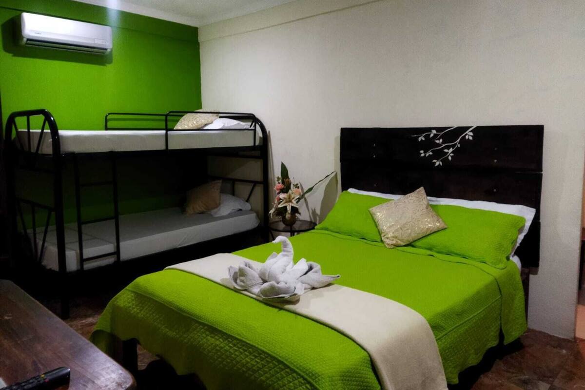 Une chambre avec des lits superposés, un lit simple avec une literie verte, une table en bois et un climatiseur fixé au mur.