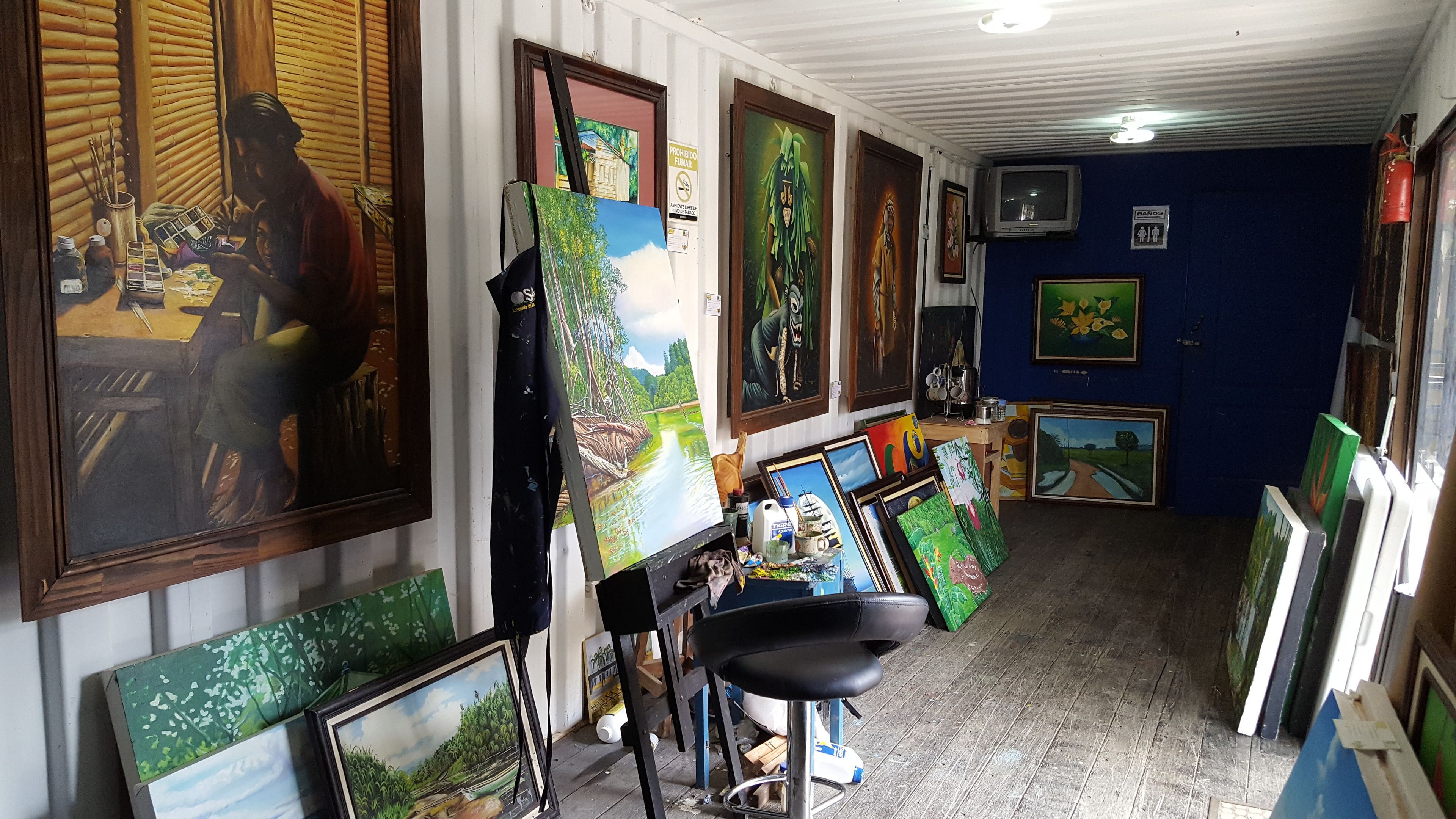 Una habitación con varias pinturas enmarcadas en las paredes, una mesa de madera con material de arte y una silla.