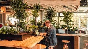 A modern workspace with wooden tables and stools, surrounded by potted plants. - ibis Budget La Rochelle Centre Gare (La Rochelle)