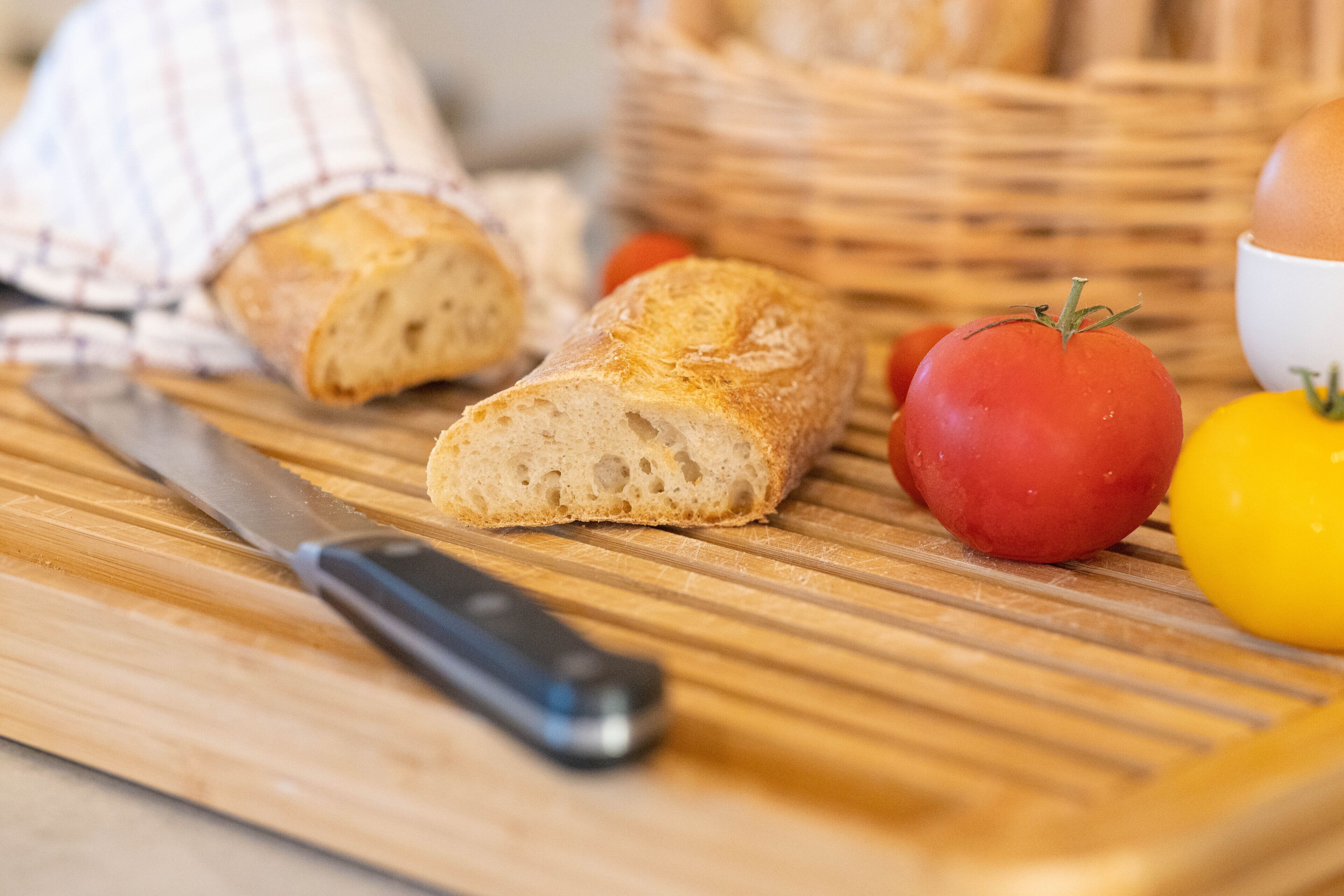 A wooden cutting board with a knife, a loaf of bread, tomatoes, and a yellow bell pepper.