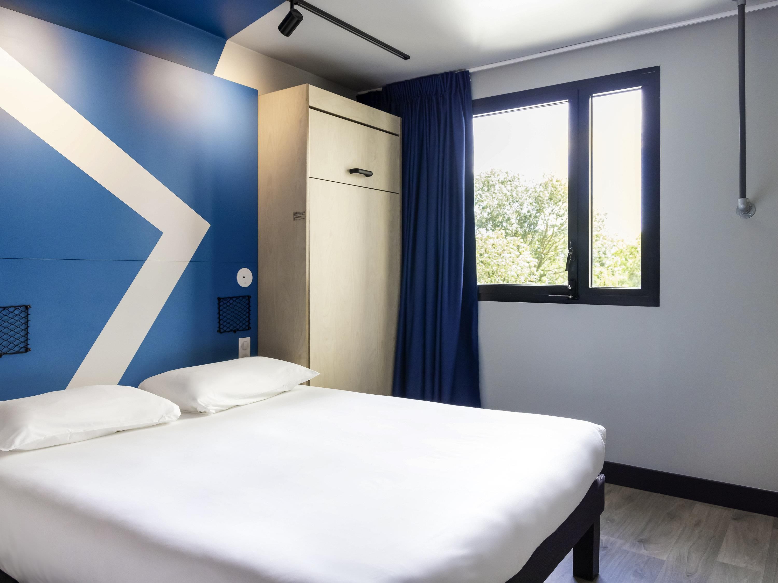 A bedroom with a bed, a wardrobe, a window with curtains, and a wall with a geometric design.