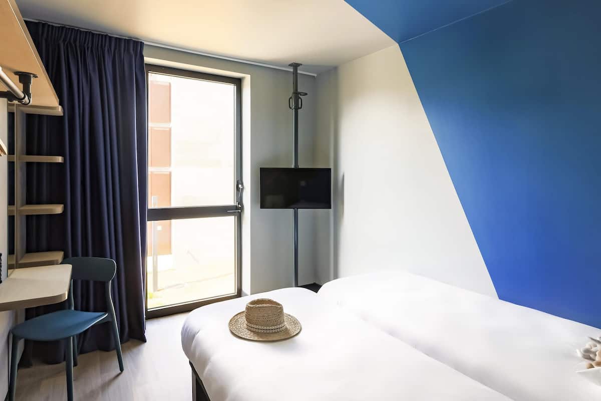 A hotel room with a bed, a desk, a chair, a TV, and a window with curtains.