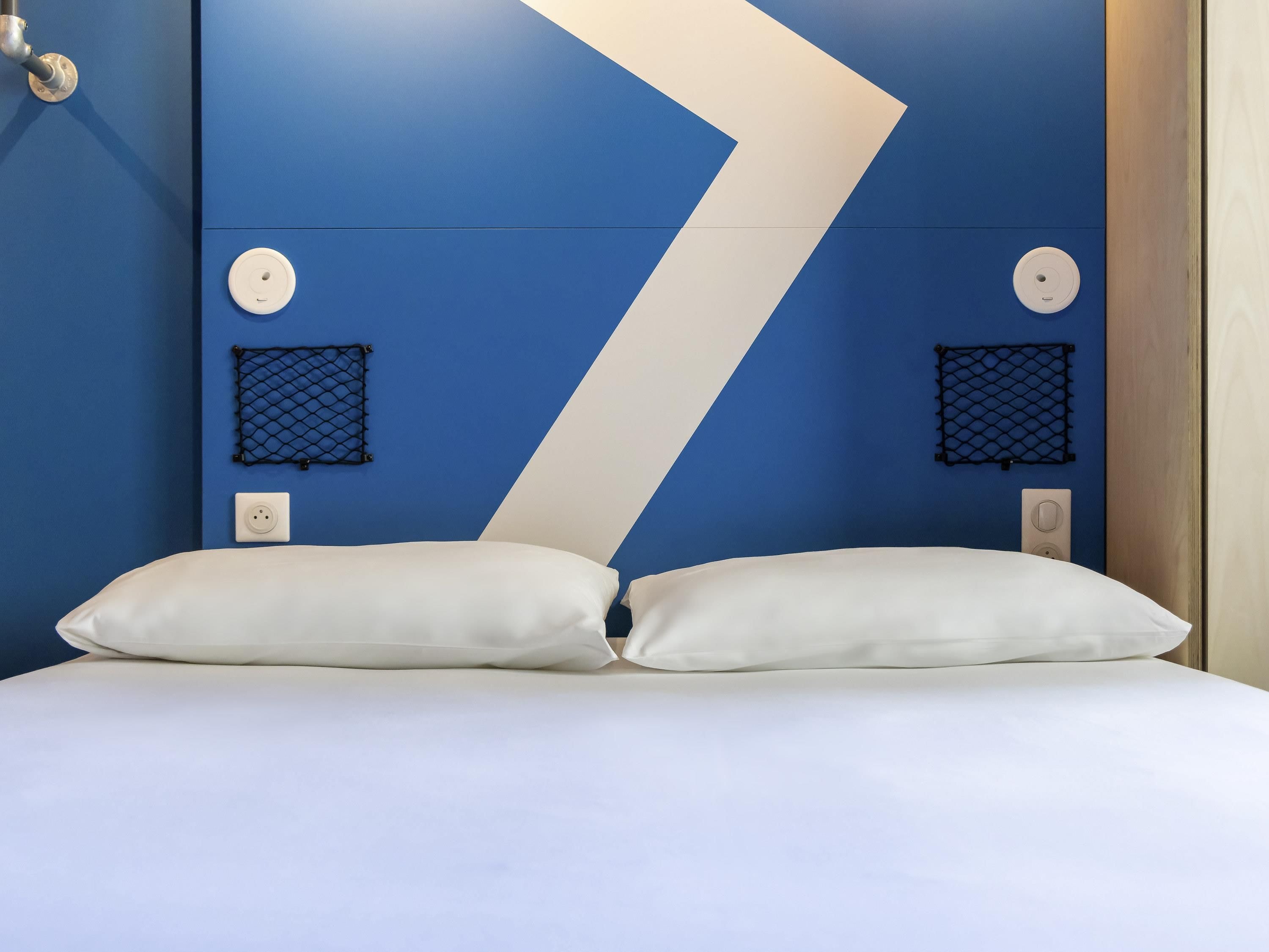 Habitación de hotel con una cama, dos almohadas blancas, una pared azul y blanca y dos paneles de malla negra pequeños.