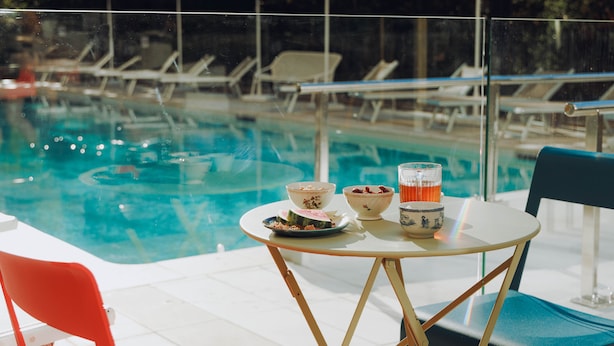 Mesa à beira da piscina com tigelas e copos, rodeada por espreguiçadeiras e guarda-sóis.