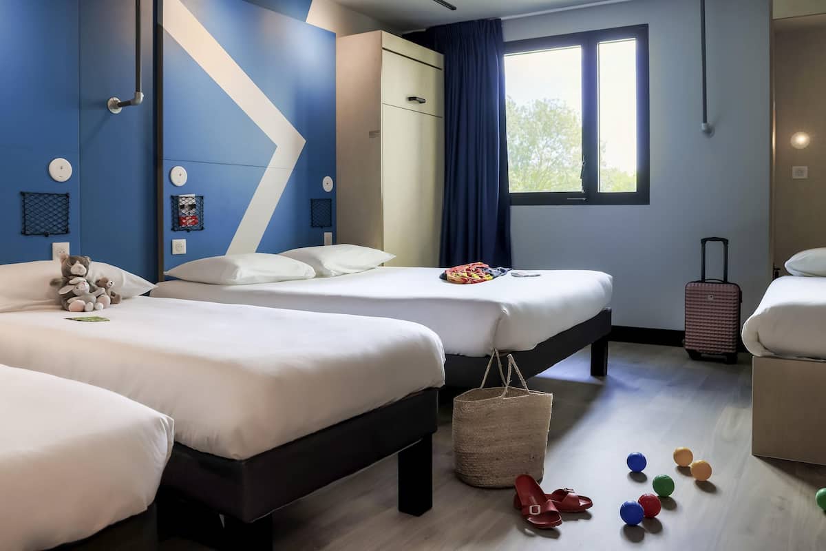 A hotel room with two beds, a large window, a suitcase, and a variety of colorful slippers on the floor.