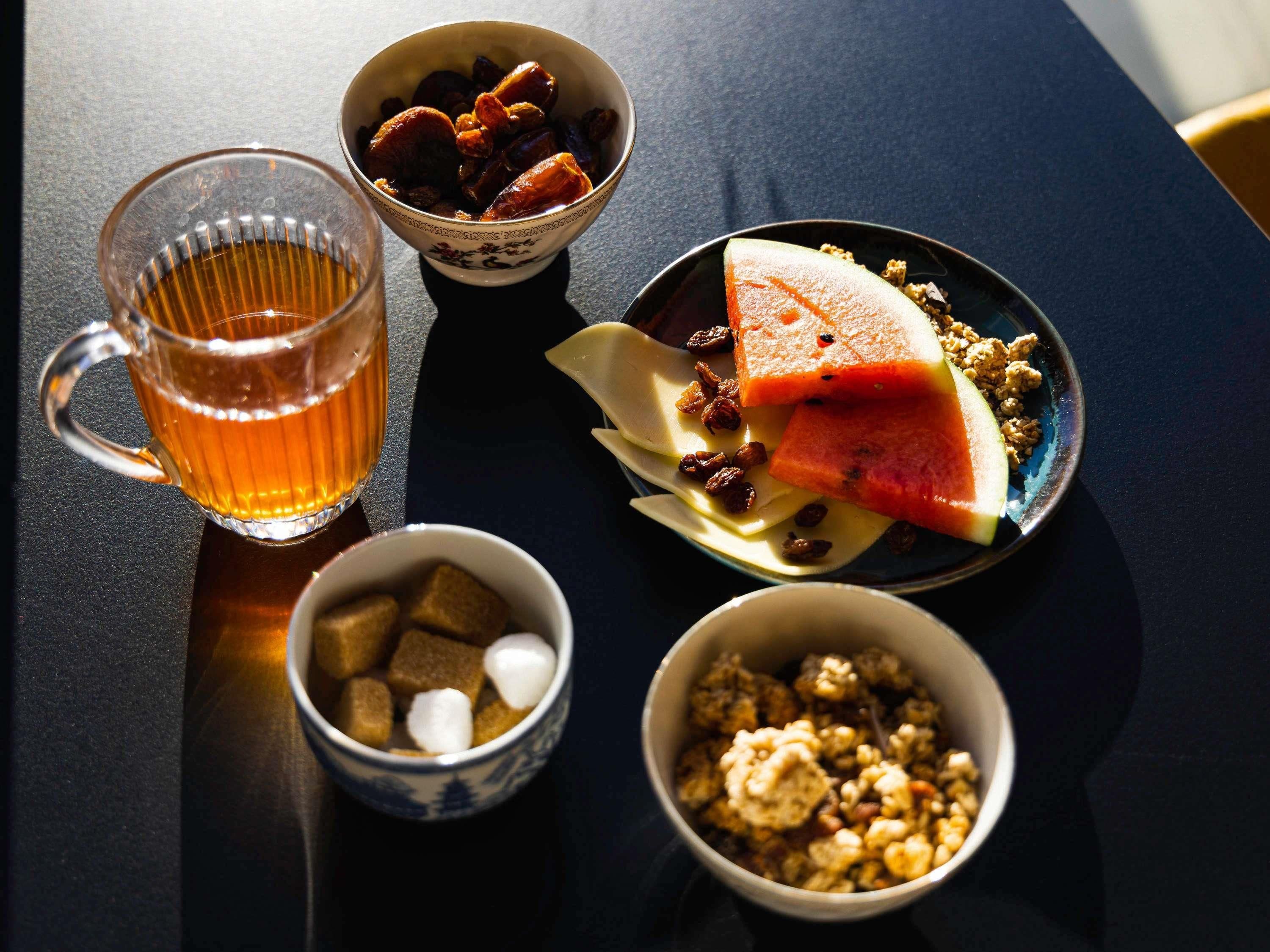Una mesa con un vaso de líquido ámbar, recipientes con frutas secas, una porción de sandía y un tazón de nueces.