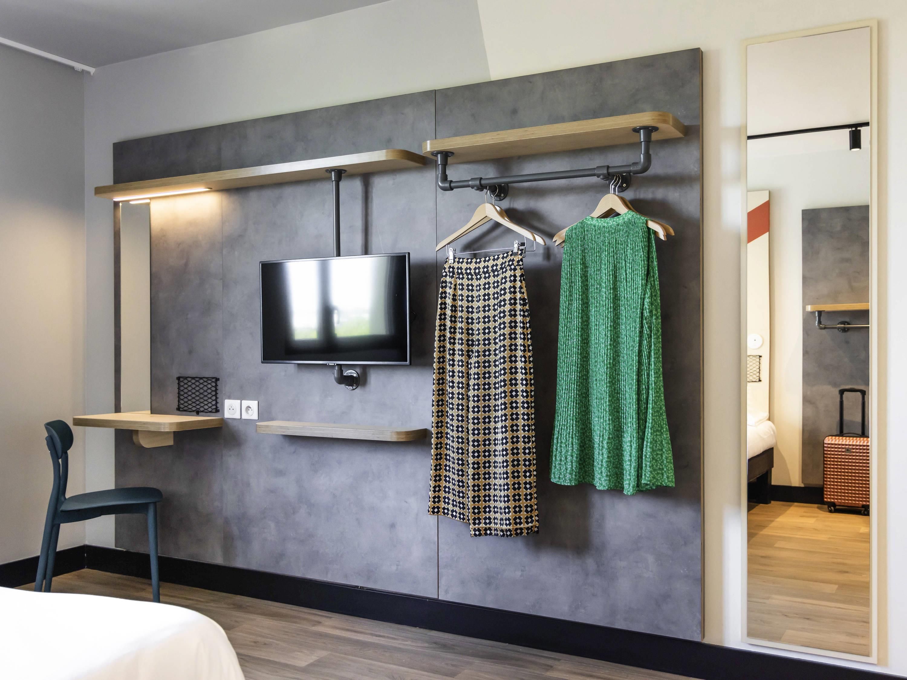 Une chambre d’hôtel dotée d’un téléviseur à écran plat fixé au mur, d’une étagère en bois et d’un portant à vêtements où sont suspendus des vêtements à motifs et de couleur verte.