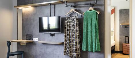 Une chambre d’hôtel dotée d’un téléviseur à écran plat fixé au mur, d’une étagère en bois et d’un portant à vêtements où sont suspendus des vêtements à motifs et de couleur verte.