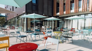 Uma área de estar ao ar livre com diversas mesas e cadeiras, guarda-sóis e um edifício moderno ao fundo.