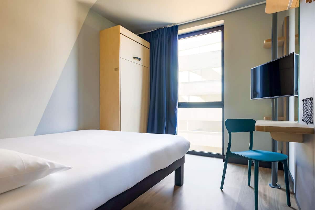 A hotel room with a bed, a desk, a chair, a TV, and a window with curtains.