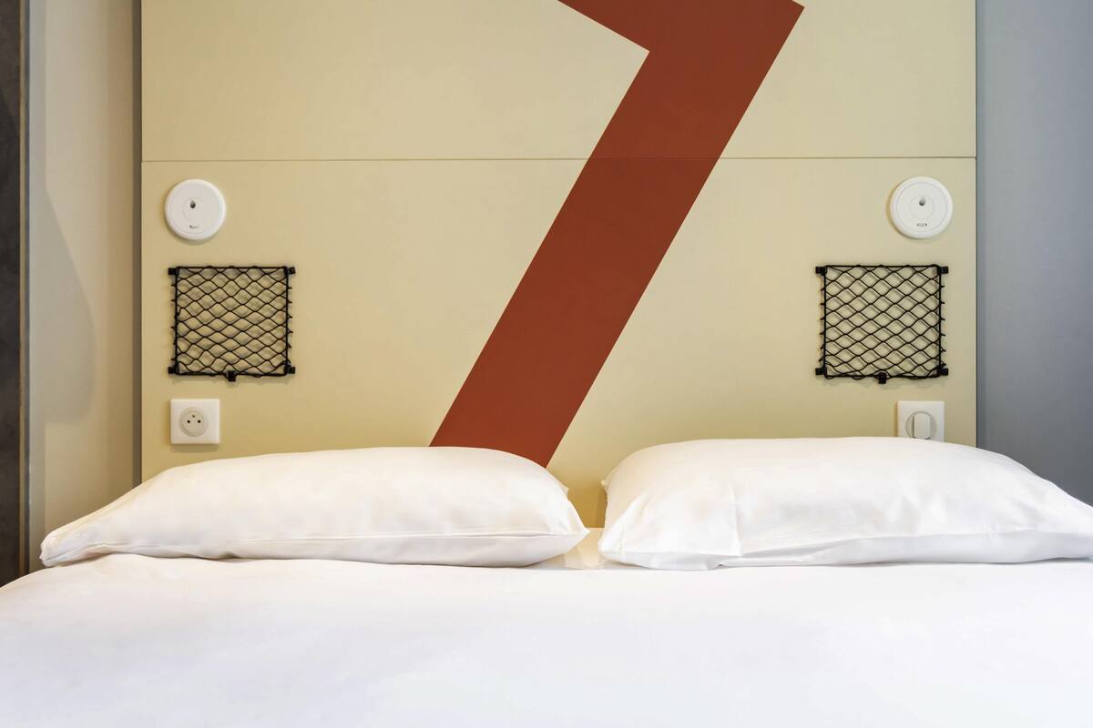 A hotel room with a large bed, two white pillows, a beige headboard, two wall-mounted power outlets, and two wall-mounted light switches.