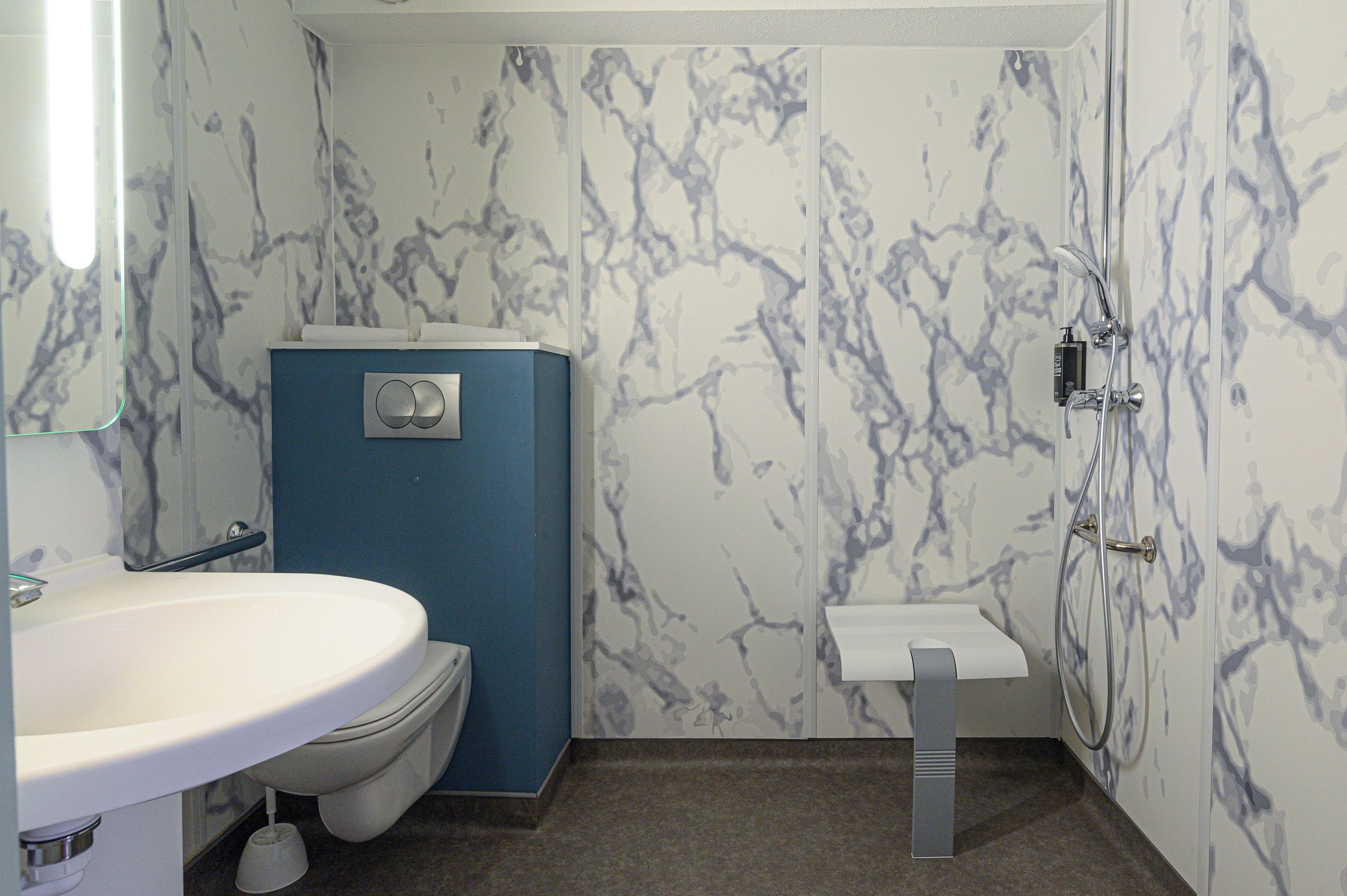 Une salle de bain avec un lavabo blanc, des toilettes bleues et une douche dont le mur est décoré de motifs en marbre.