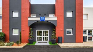 Un hotel moderno con fachada roja y gris, un letrero azul que indica que es un hotel ibis budget, y una entrada despejada con plantas en macetas.