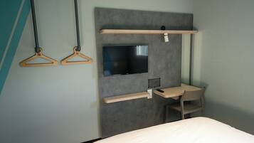 A hotel room with a bed, a desk, a chair, a television mounted on a grey wall, and two wooden hangers.