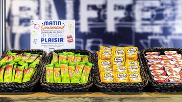 A display of assorted packaged goods in baskets, including items labeled 'MEL - HONEY' and 'MEL - HONEY + JEL,' with a sign promoting 'MATIN - Gourmand' and 'LA TOUCHE PLAISIR.'