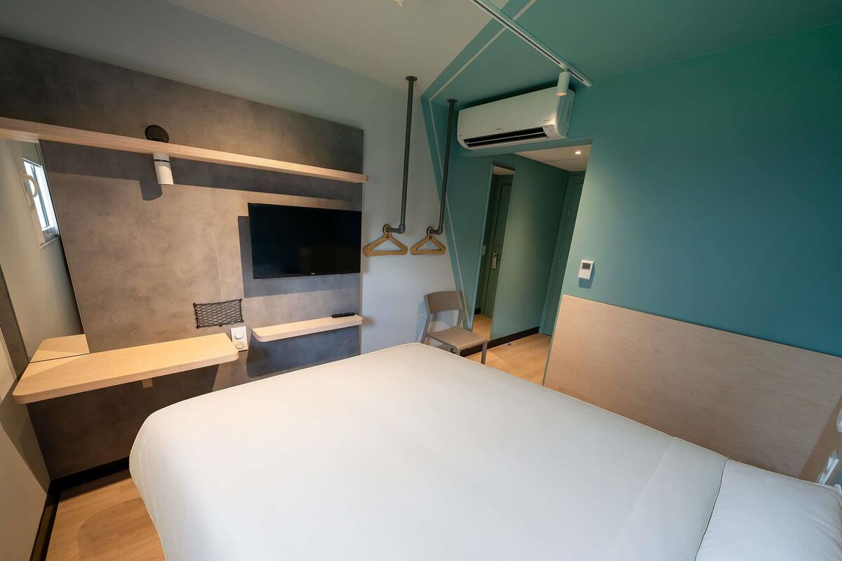Una habitación de hotel con una cama, un televisor montado en la pared, un pequeño rincón de descanso y un equipo de aire acondicionado.