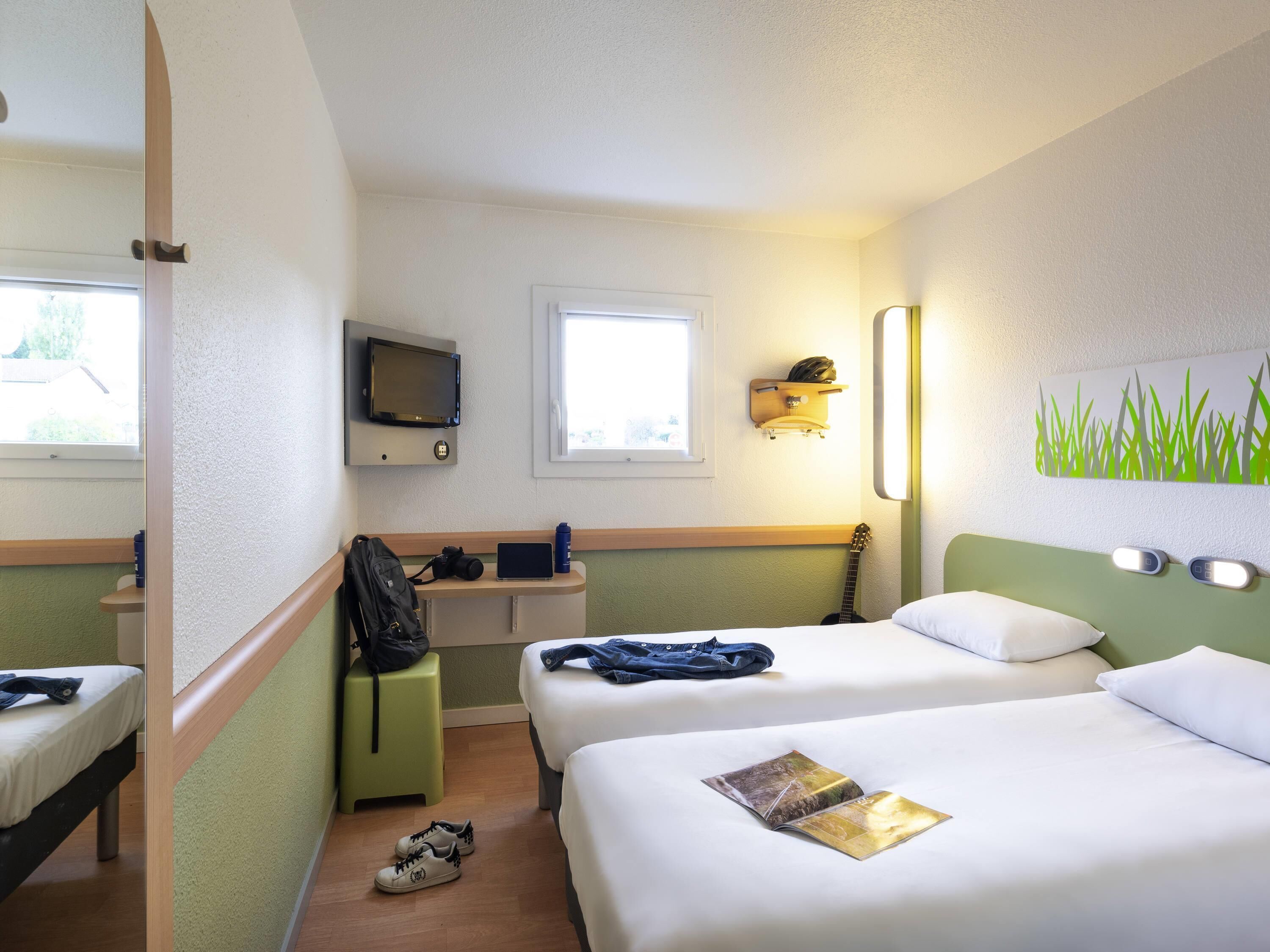 A hotel room with two beds, a desk with a computer, a TV, a window, and a wall-mounted shelf.