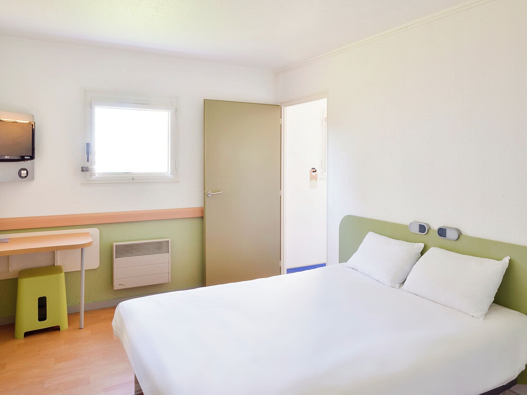 Una camera d'albergo con un letto, una scrivania, una finestra e una TV a parete.