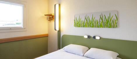 Una habitación de hotel con una cama, un ventanal, una lámpara fijada a la pared y un cuadro con césped.