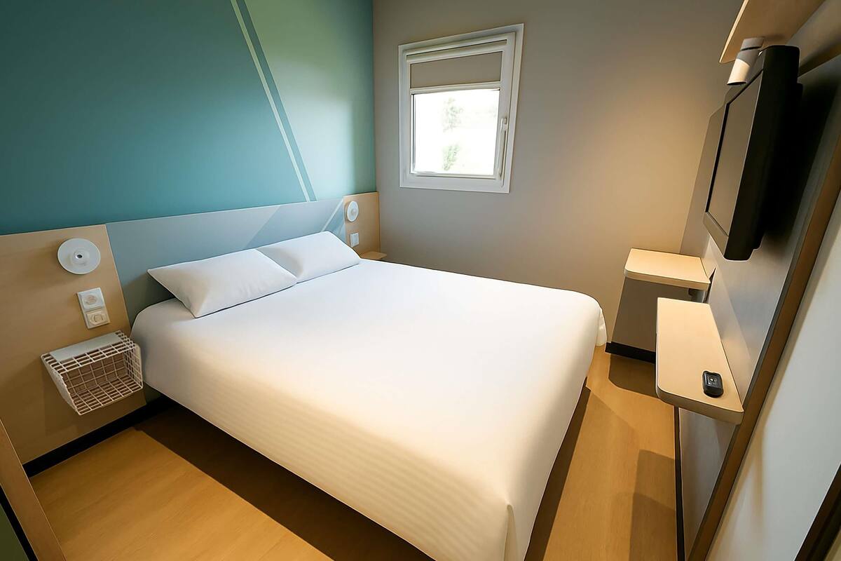 Una cama bien tendida en una habitación con ventana, una mesita de noche con control remoto y un televisor montado en la pared.