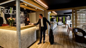 Zona de recepción con mostrador de madera, un hombre con traje y maleta, y un interior moderno con piso de madera y detalles verdes.