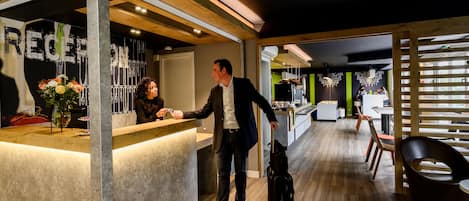 Zona de recepción con mostrador de madera, un hombre con traje y maleta, y un interior moderno con piso de madera y detalles verdes.
