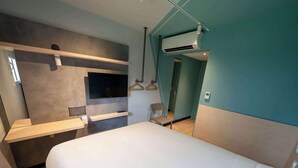 Una habitación de hotel moderna con una cama grande, un televisor montado en la pared, un aire acondicionado instalado y una pequeña zona de estar con una silla y un perchero.