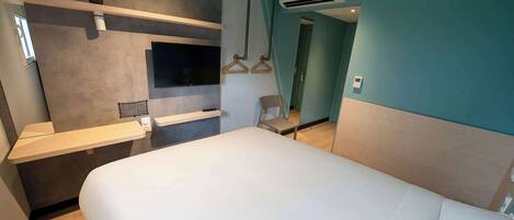 Une chambre d’hôtel moderne dotée d’un grand lit, d’un téléviseur fixé au mur, d’un climatiseur mural et d’un petit coin salon avec une chaise et un porte-manteau suspendu.
