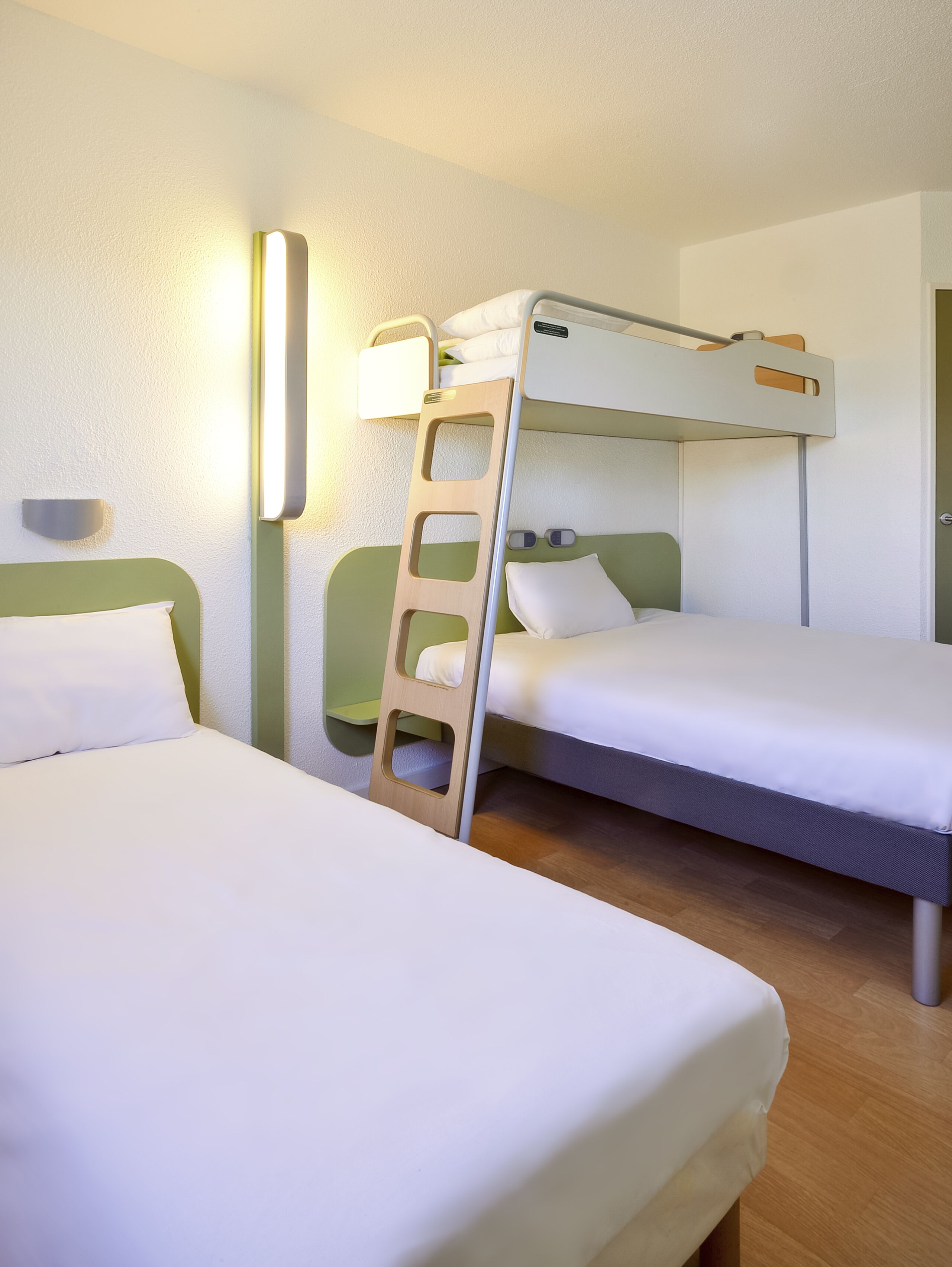 A hotel room with two single beds, a wooden ladder, a wall-mounted light, and a door.