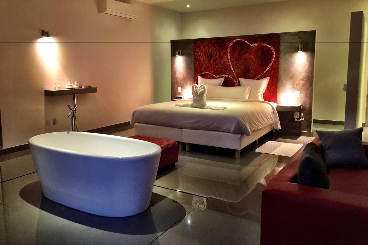 Une chambre à coucher moderne avec une grande baignoire, un lit doté d’une tête de lit en forme de cœur et deux lampes de chevet.