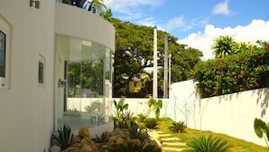 Una casa moderna con fachada de vidrio, un camino de piedra y un jardín exuberante con diversas plantas y un pequeño estanque.