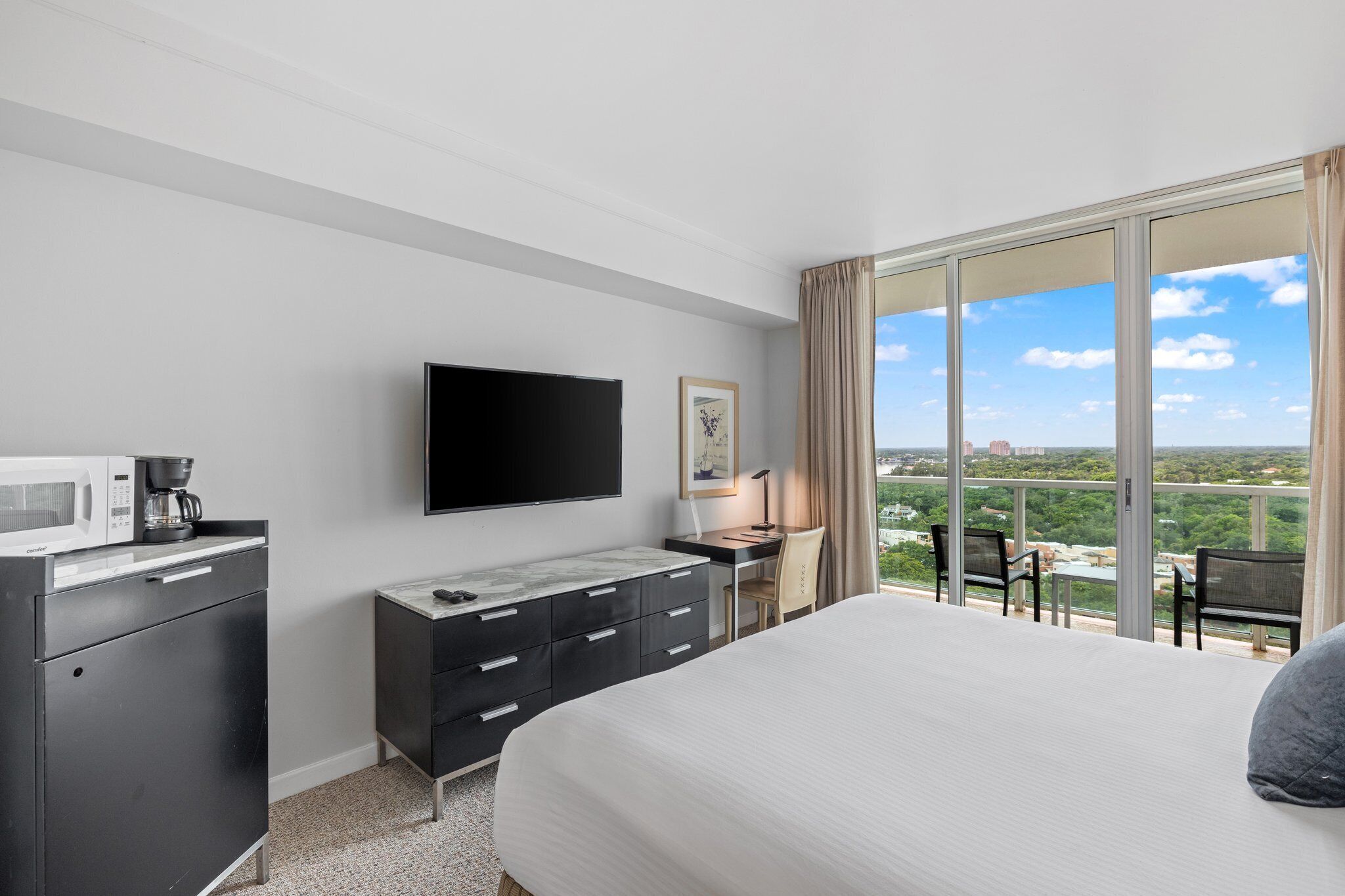 A hotel room with a large bed, a flat-screen TV, a desk with a chair, a coffee maker, and a view of the city through large windows.