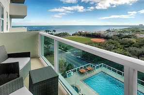 Un balcón con mobiliario de mimbre, una piscina y vistas al mar y al paisaje urbano.