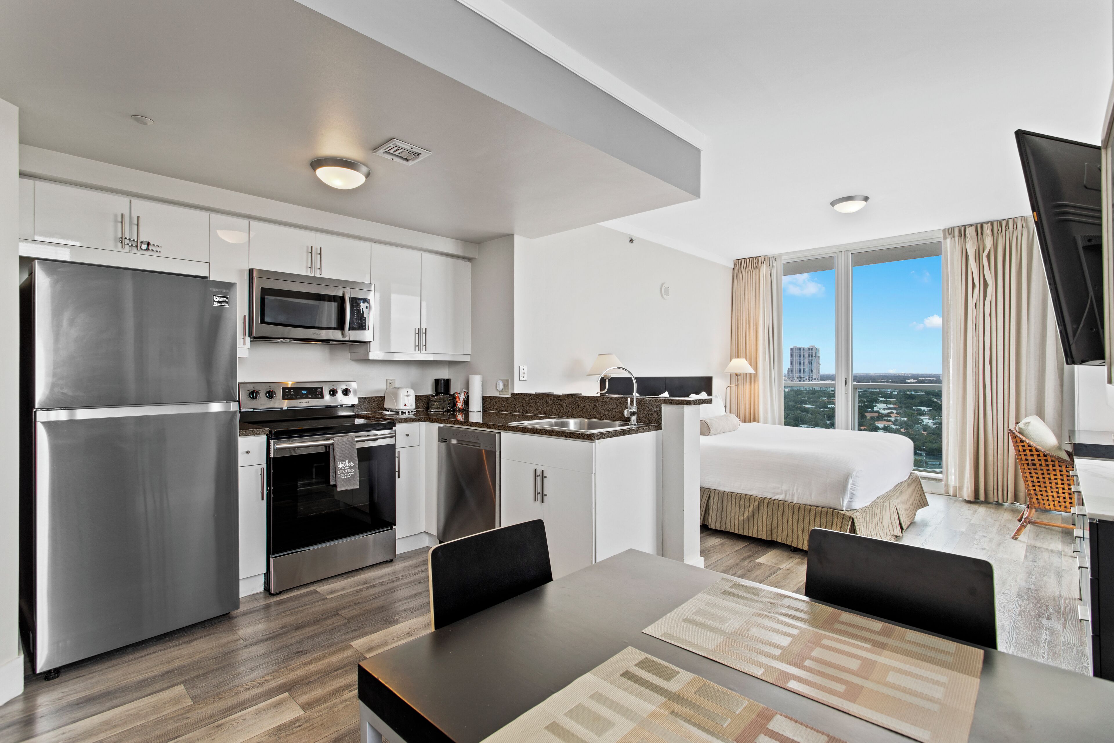 A modern hotel room with a kitchenette, a bed, a TV, and a city view.