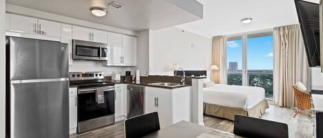 A modern hotel room with a kitchenette, a bed, a TV, and a city view.